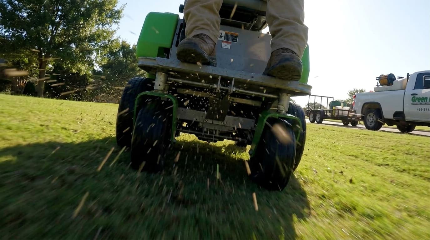 A Green Lawn Fertilizing technician on a stand-on spreader applying treatment across a lawn, with a branded truck and trailer in the background